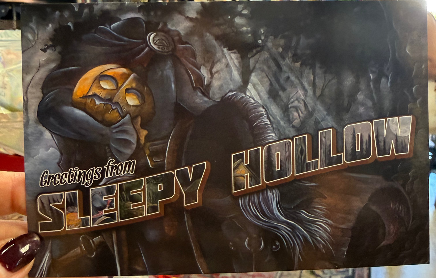 a printed card or art piece featuring a dark illustrated scene with a ghostly figure and the text 'Greetings from Sleepy Hollow' in large stylized letters. 